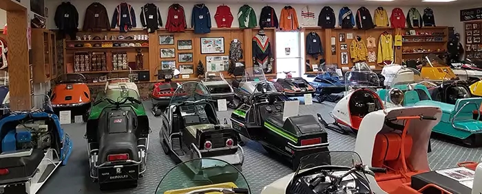 Room with three rows of vintage snowmobiles, snowmobile jackets hung along the back wall, and shelves of snowmobile memorabilia. 