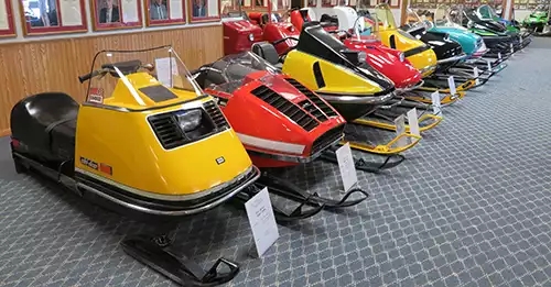 A dozen vintage snowmobiles in a row, starting with an old yellow skidoo. 