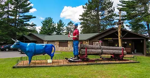 Foreground is a blue ox pulling a flatdeck of logs with Paul Bunyan standing on it holding an ax. Museum in the background. 
