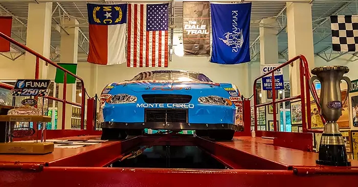 blue Monte Carlo car on a lift, with flags hung above it. A trophy sits in the right corner. 